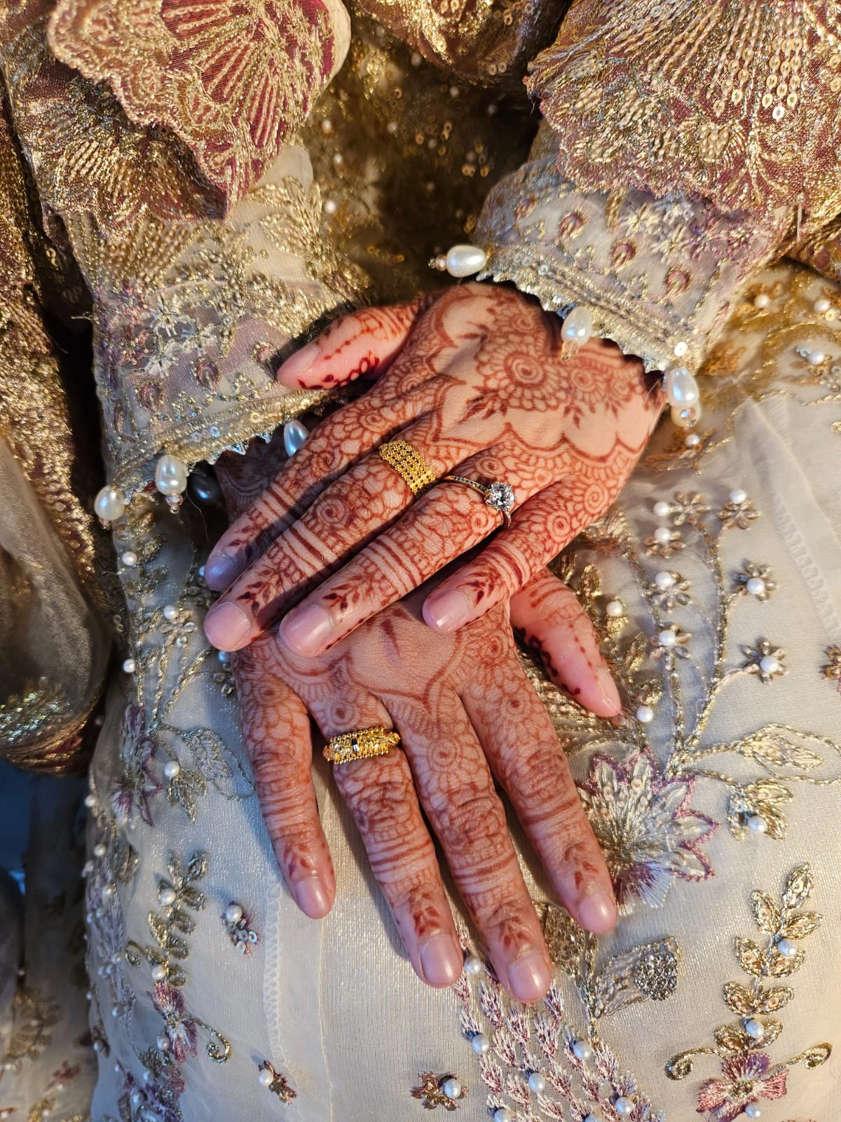 Floral mehndi on the backs of two hands, with gold rings and embroidered bridal sleeves